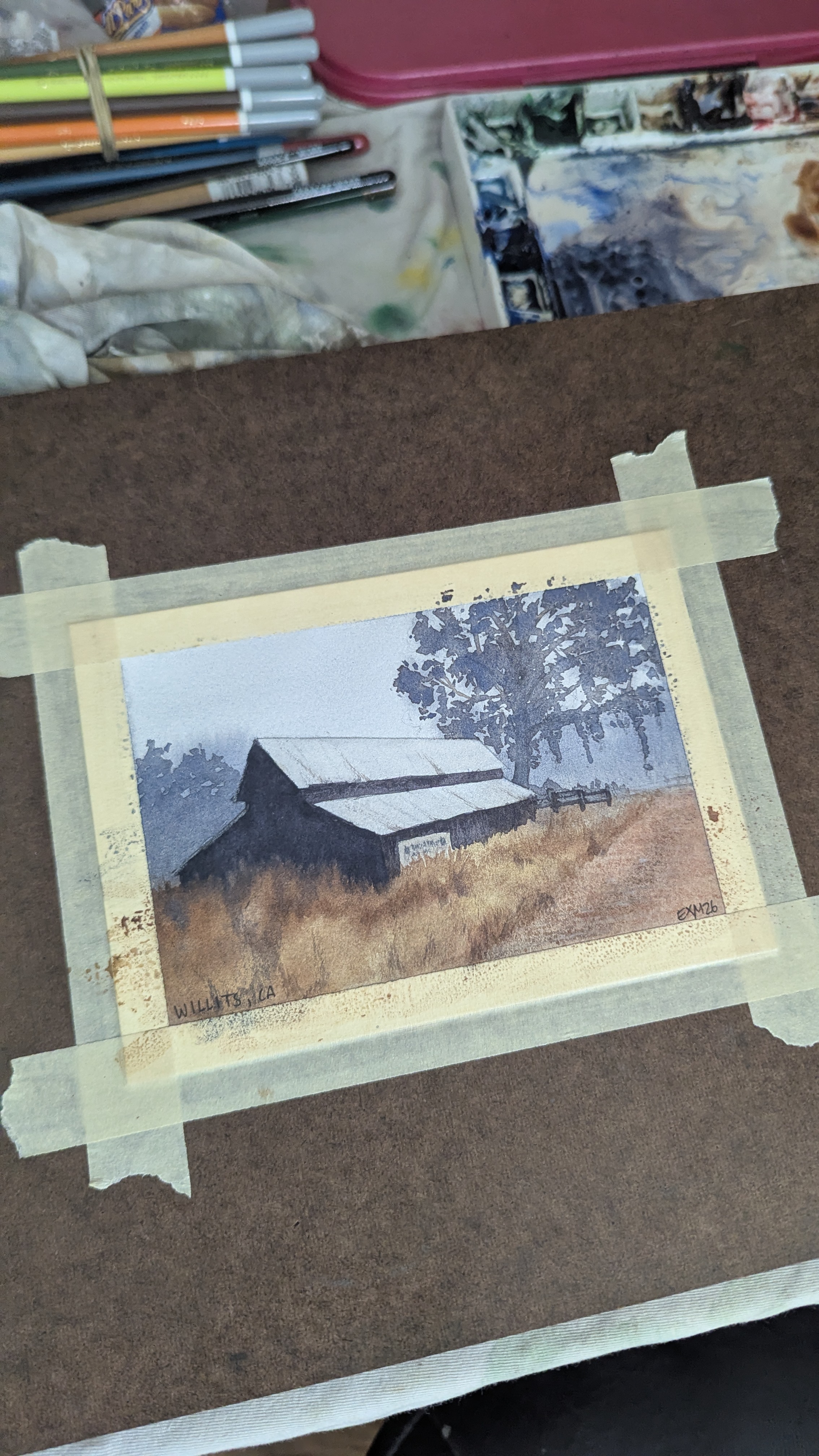 Watercolor painting of a barn in the morning fog in Willits, CA sitting on a an artist's desk.